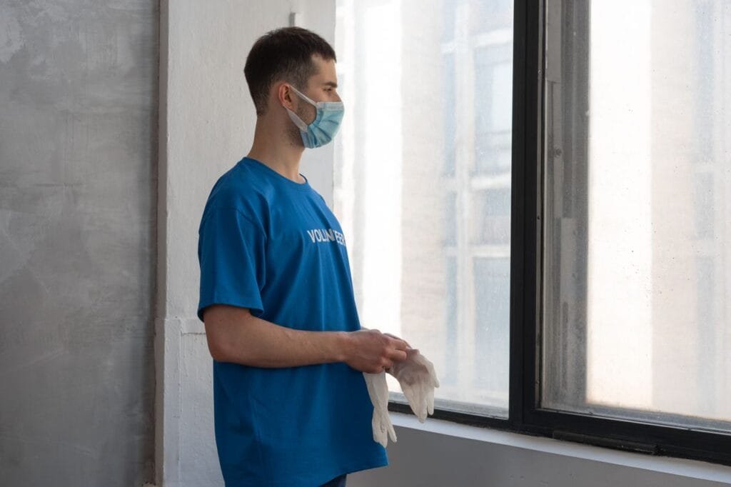 A male volunteer wearing a face mask stands by a window indoors, holding gloves.